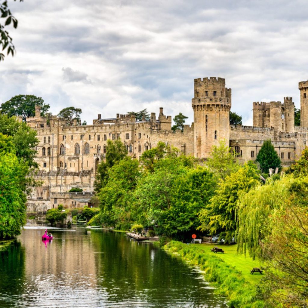 Warwick castle from the river with trees surrounding the river banks. Visiting here is one of the things to do in England